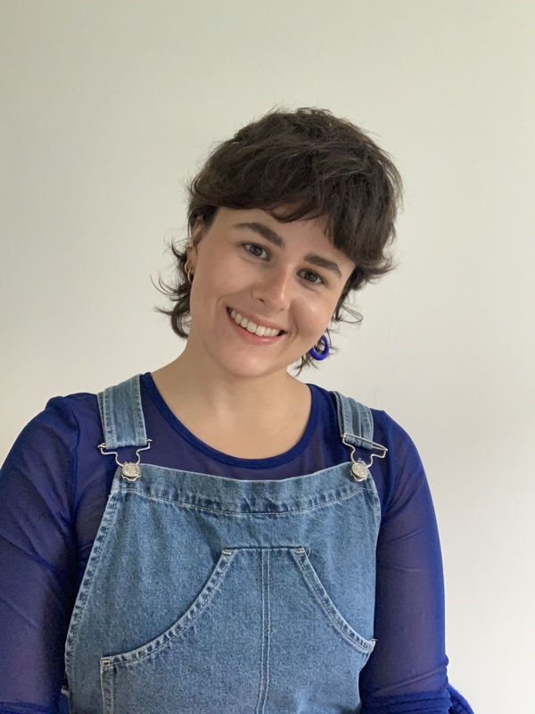 Adèle Gonzalez en salopette bleue, souriante la tête penchée, sur un fond crème.